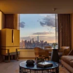 Modern furnished corporate apartment interior with sofa, workspace, and large window overlooking the New York City skyline at sunset.